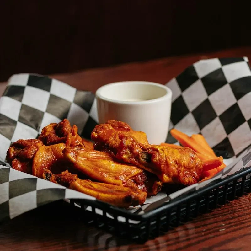 Close-up of buffalo wings, carrots, and creamy dip in a checkered basket.
