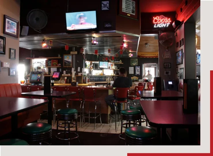 Dimly lit bar interior with counter, bottles, screens, stools, and neon sign.