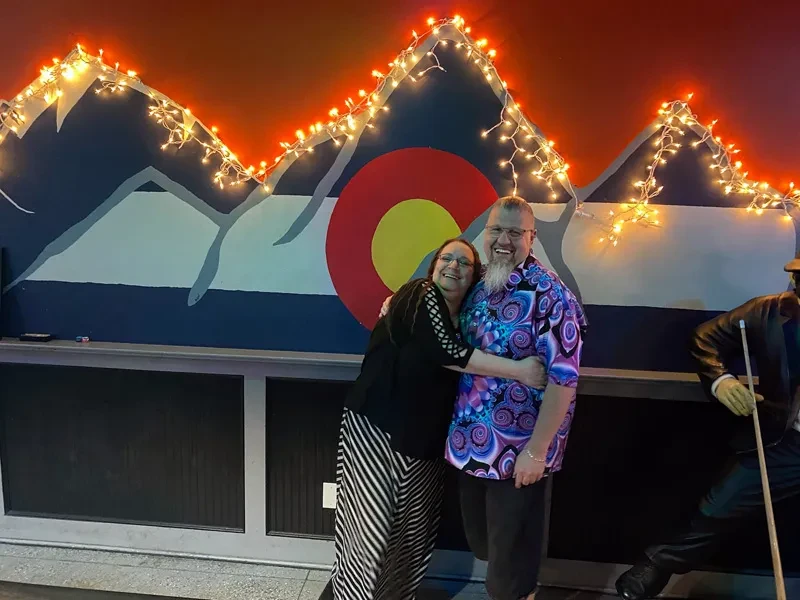 Smiling man and woman embrace before a vibrant lit Colorado mountain mural.