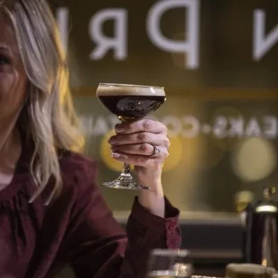 Hand holding a dark, foamy cocktail in a coupe glass.