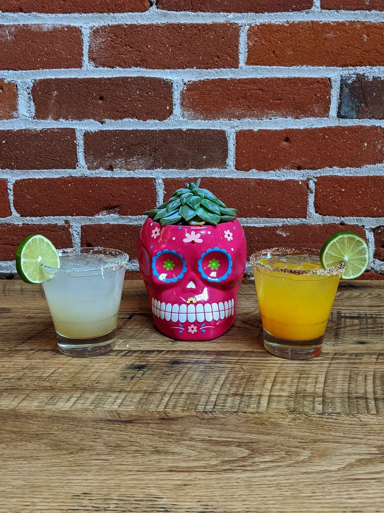 Two garnished drinks, pink sugar skull planter on rustic wood, brick background.