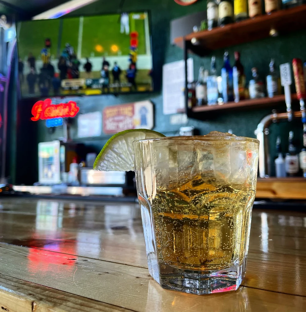 Golden drink with lime, ice on bar counter, blurred sports.