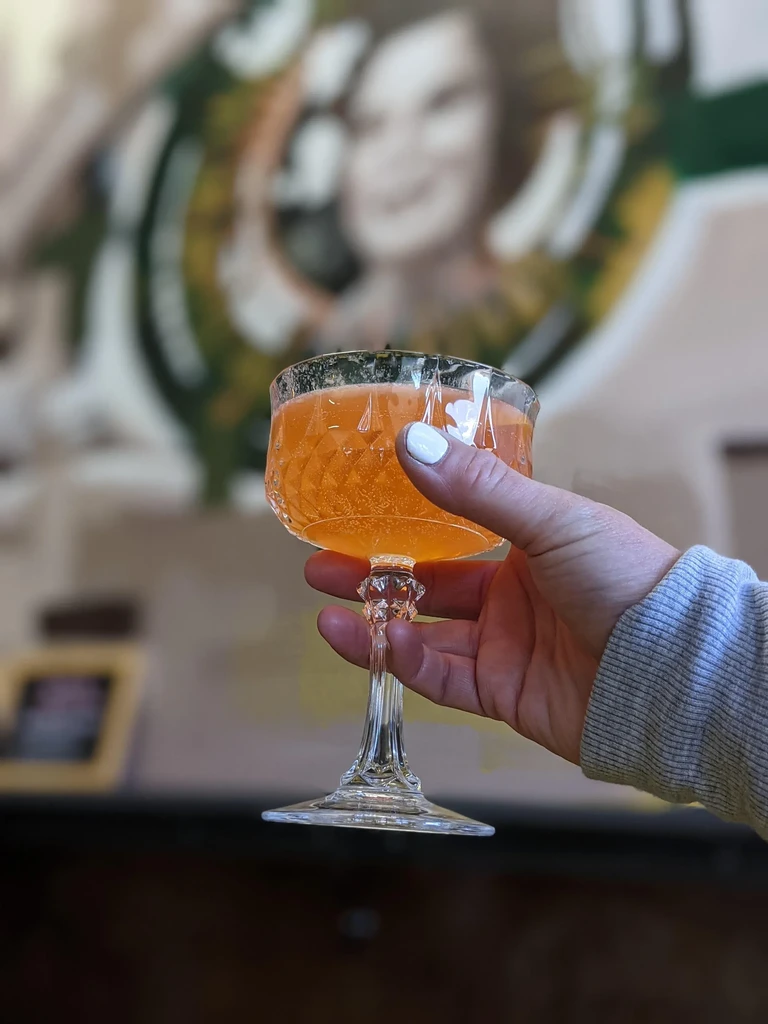 Hand holds orange cocktail in textured glass; blurred art background.