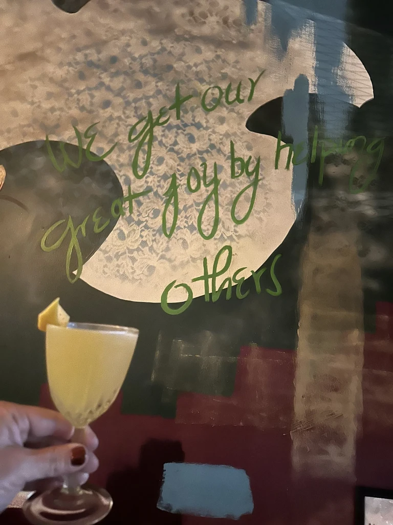 Manicured hand holds a lemon cocktail; green script wall quote.