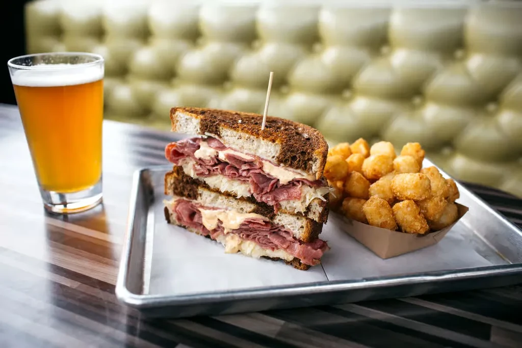 A Reuben sandwich, tater tots, and beer on a metal tray.