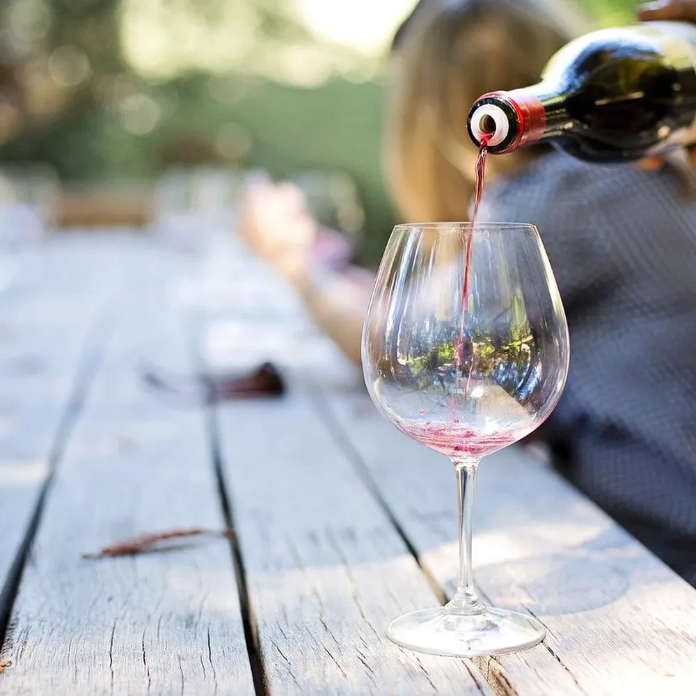 Red wine pours from bottle into glass on rustic outdoor table.