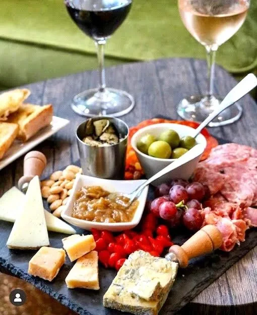 Charcuterie board with cheeses, meats, grapes, olives, and two wine glasses.