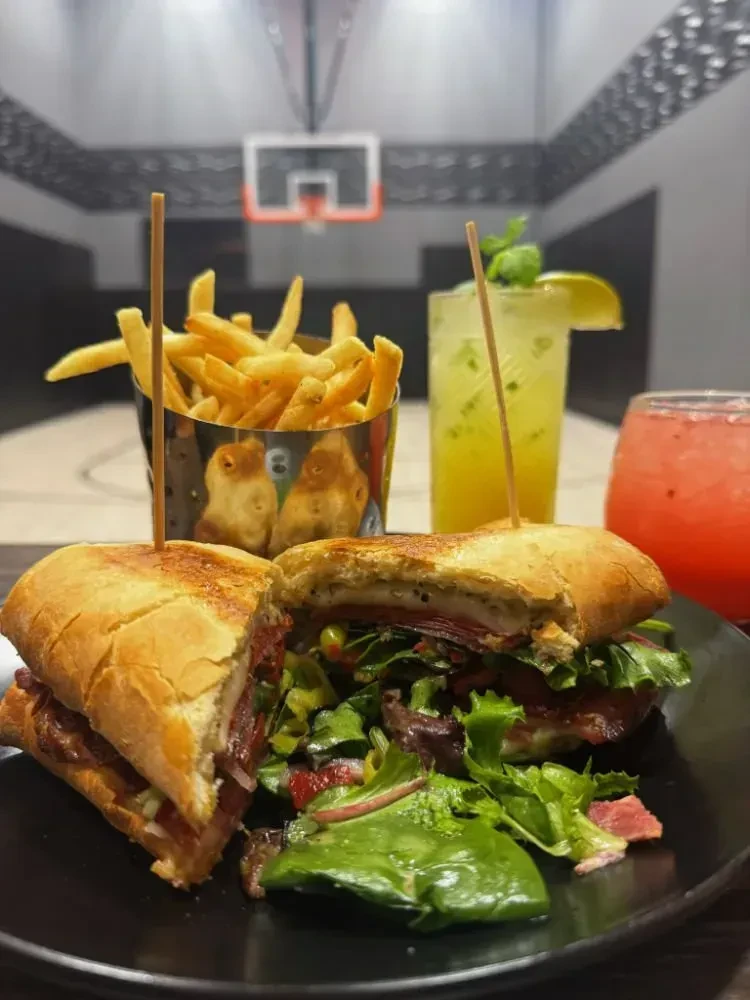 Artisanal sandwich, fries, salad, and drinks on a blurred basketball court.