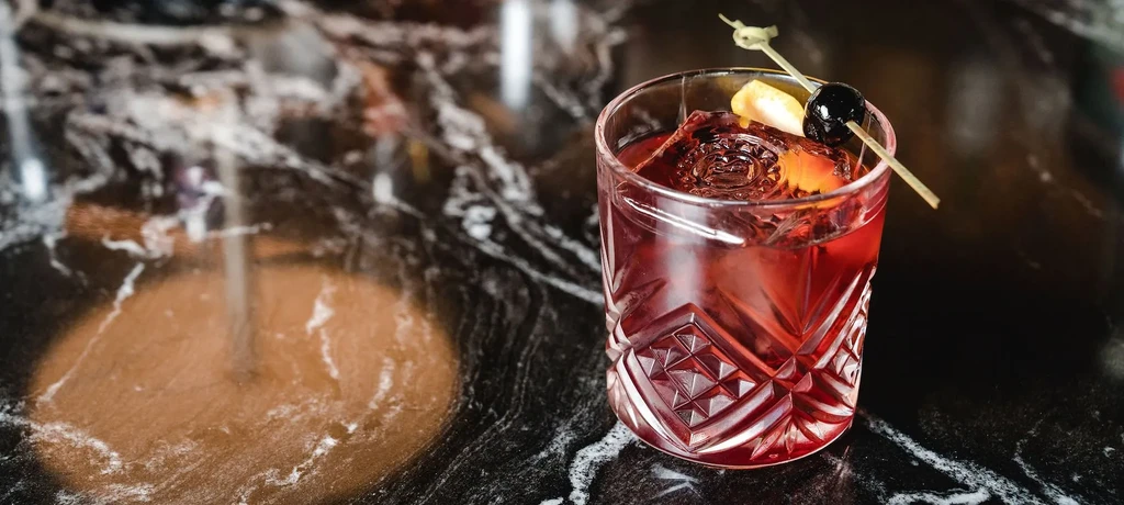 Reddish-amber cocktail with sphere ice, cherry, and citrus on dark marble.
