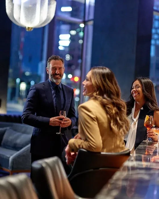Three people socialize in a modern, upscale city bar at night.