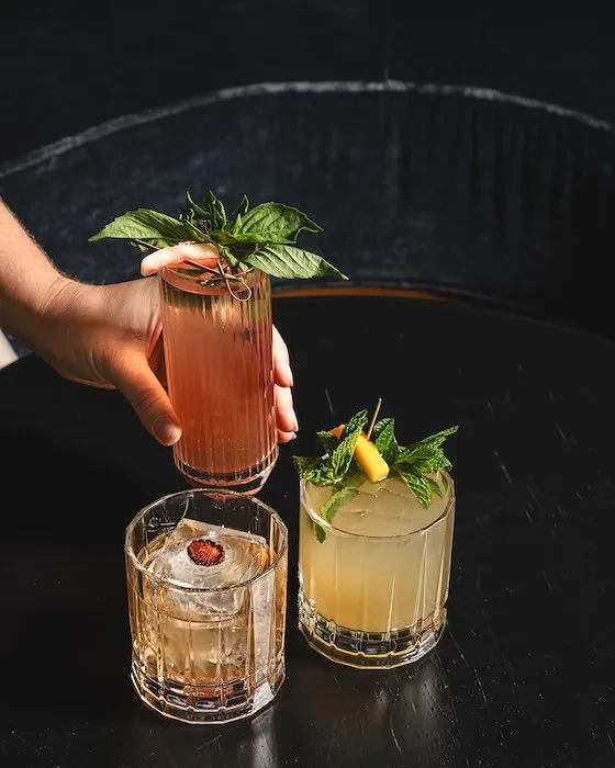 Three elaborately garnished cocktails on a dark tabletop, one held by a hand.