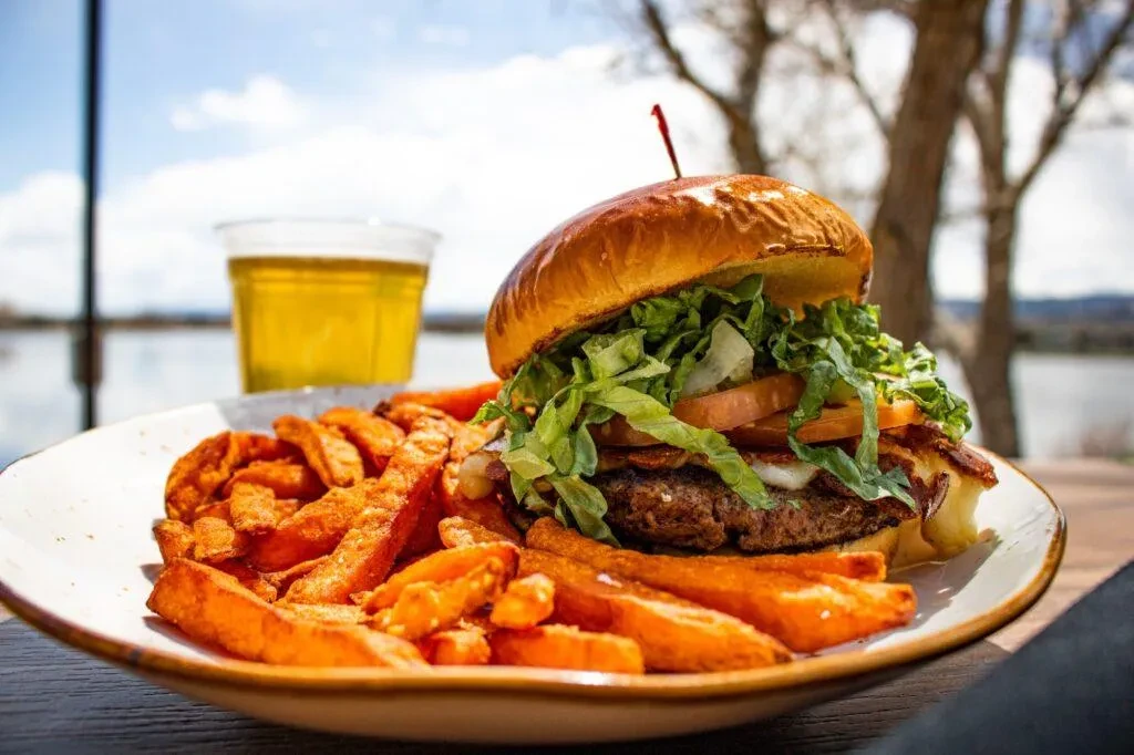 Gourmet hamburger and sweet potato fries outdoors by water.