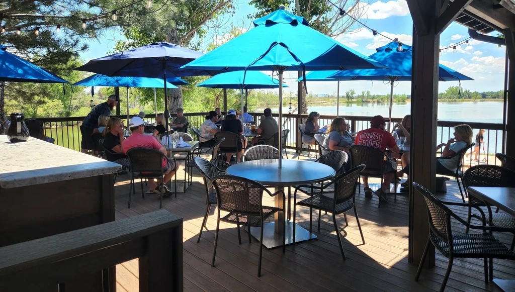 Busy restaurant patio overlooking a lake, with patrons under blue umbrellas.