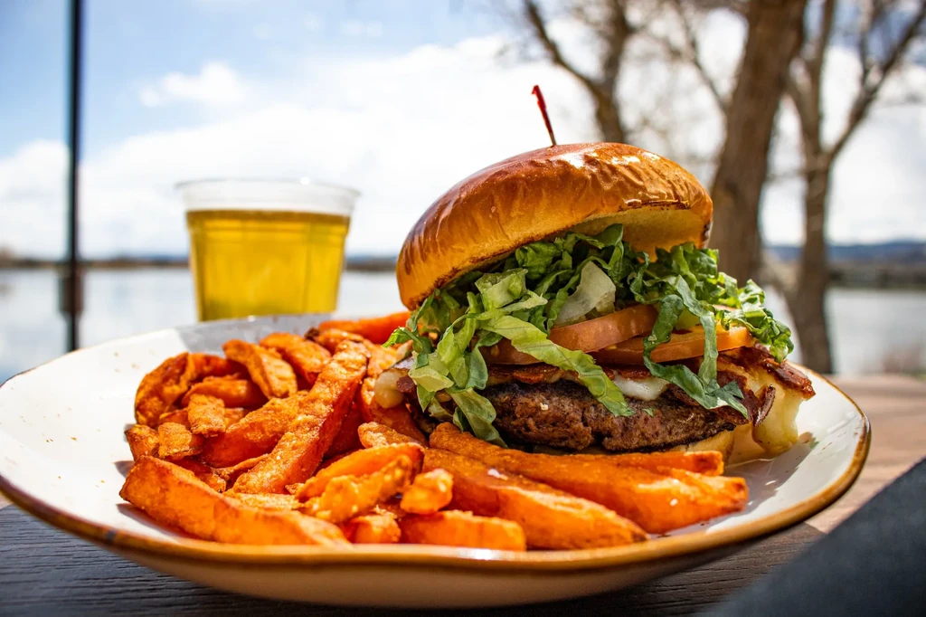 Close-up burger, sweet potato fries, and beer outdoors by a lake.