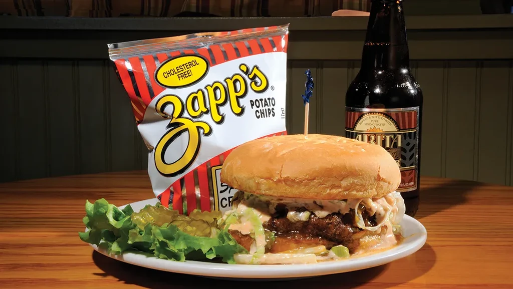 Burger, Zapp's chips, and Abita beer served on a rustic table.