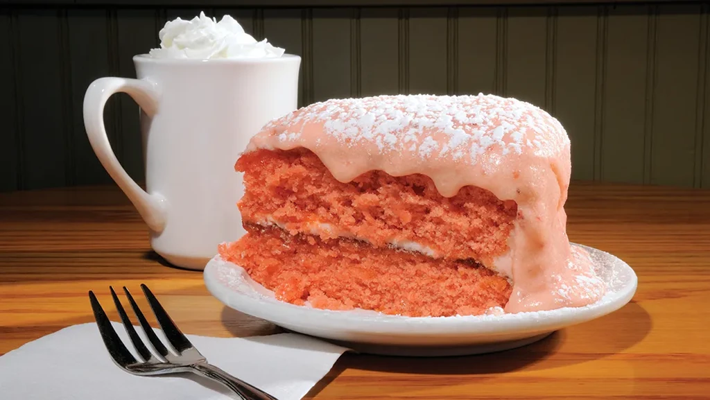 Pink glazed cake slice, whipped cream mug, fork on wooden table.