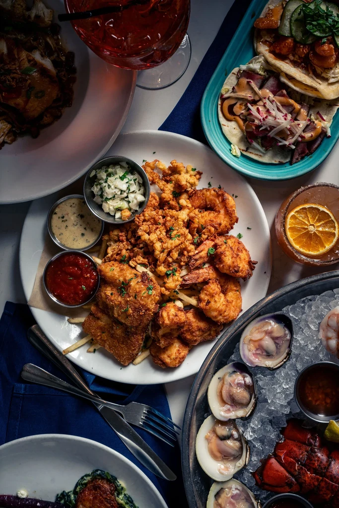 Diverse fried seafood, tacos, raw bar, and cocktails on a table.