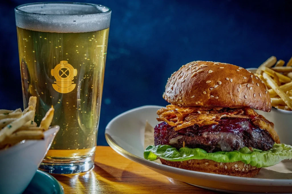 Close-up of a burger, french fries, and cold beer with a diving helmet logo.