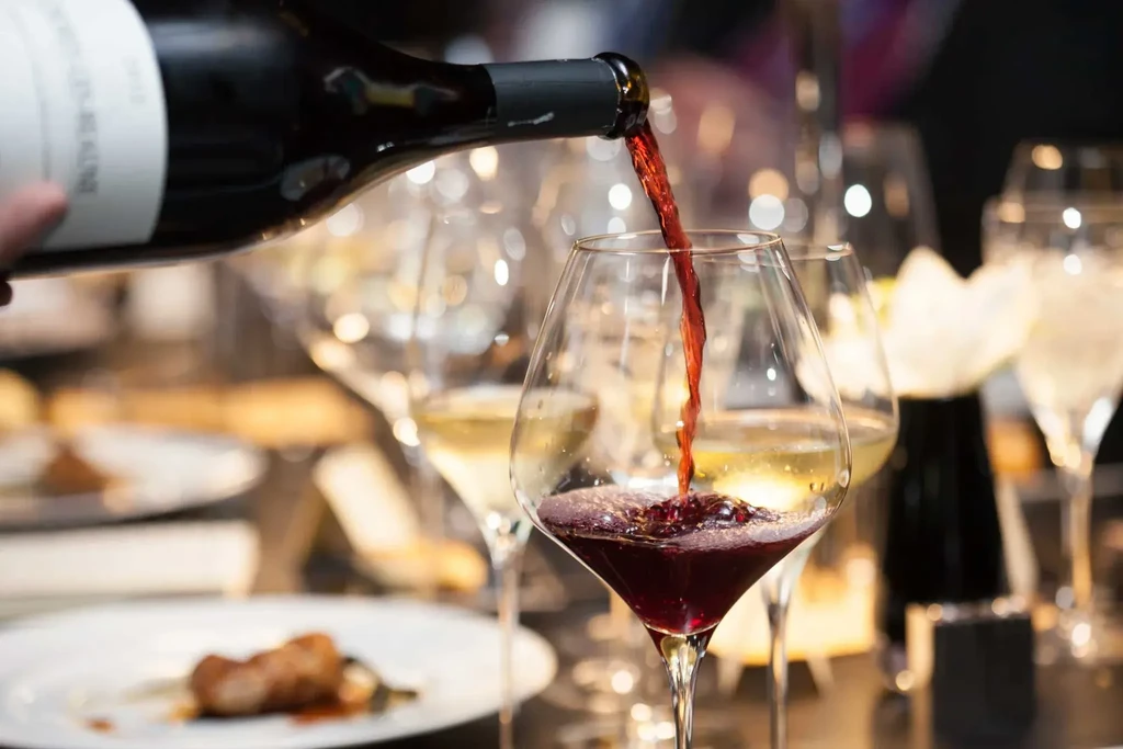 Hand pouring red wine into a glass; blurred background of food and drinks.