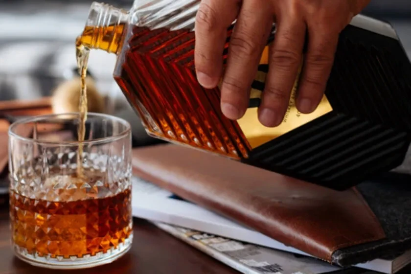 Hand pouring amber liquid from patterned bottle into whiskey glass.
