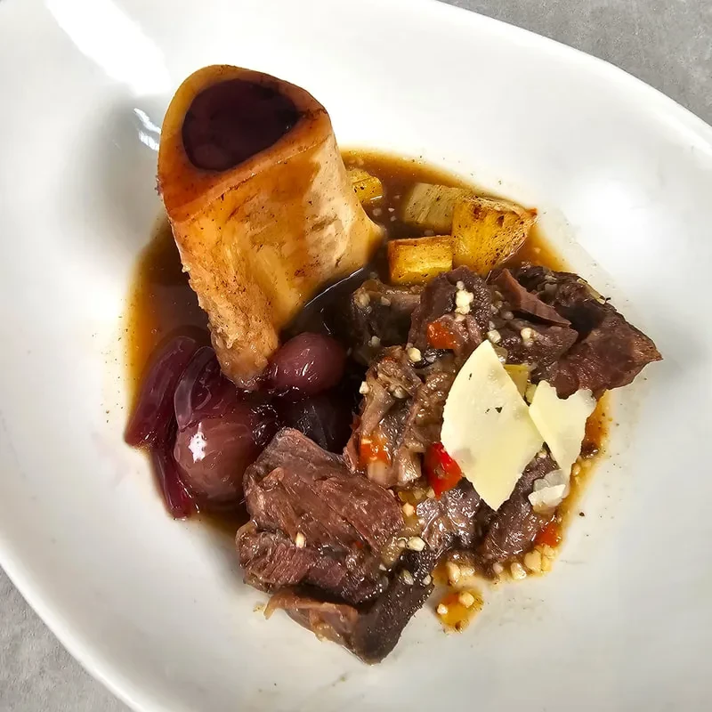 Hearty braised meat, marrow bone, vegetables, and cheese on a white plate.