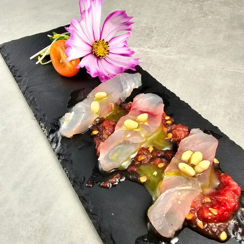 Plated white fish, red sauce, pine nuts, and flower on black slate.