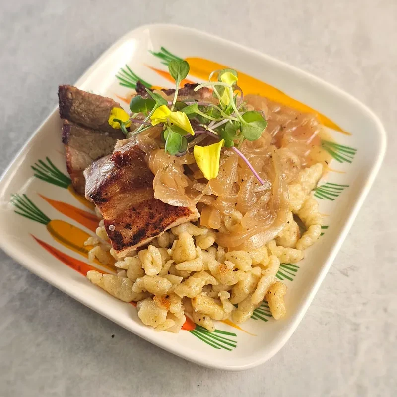 Plated seared meat, caramelized onions, spaetzle, microgreens, and edible flowers.