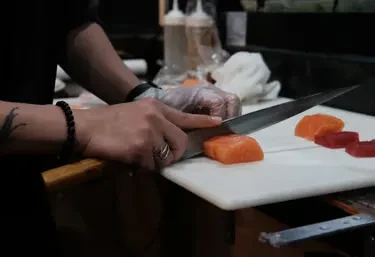 Hands, one gloved, slicing salmon sashimi with a chef's knife.