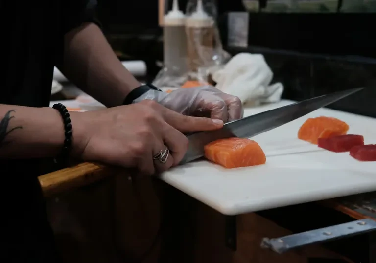 Gloved hands slicing orange salmon with a knife on a white board.