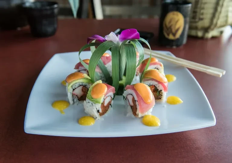 Sushi rolls topped with fish, avocado, and orchid on white plate.