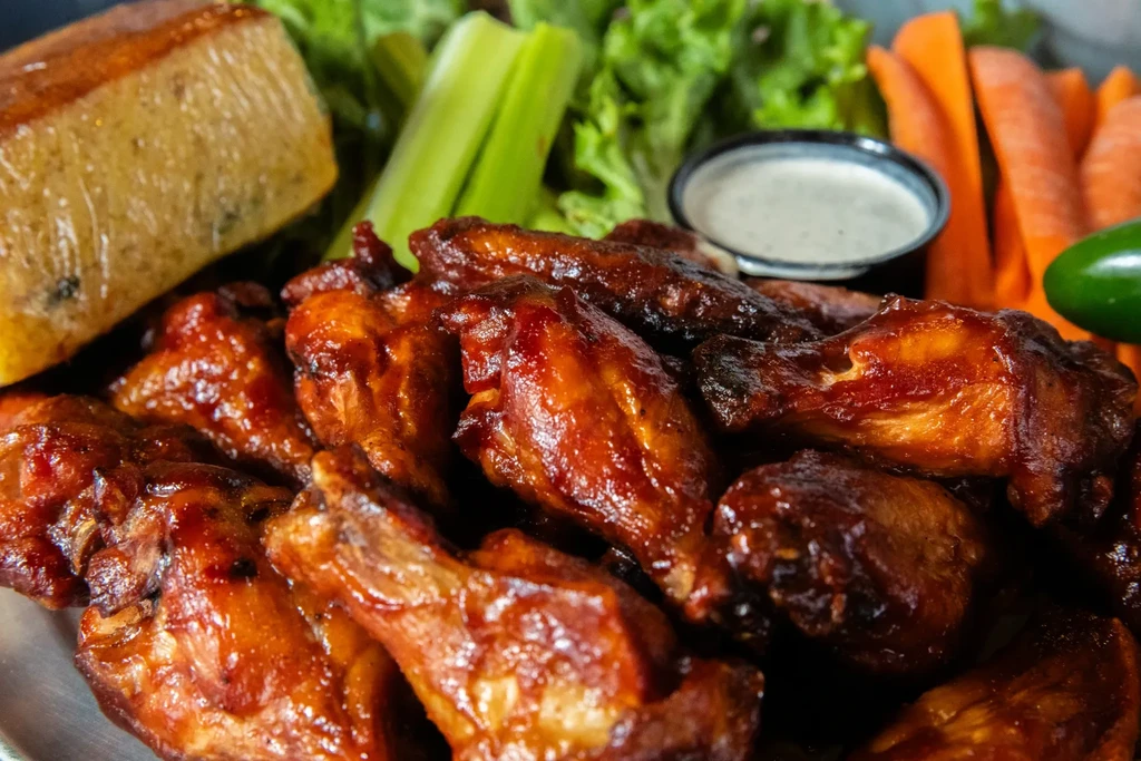 Close-up: Glazed chicken wings, cornbread, vegetables, and creamy dip.
