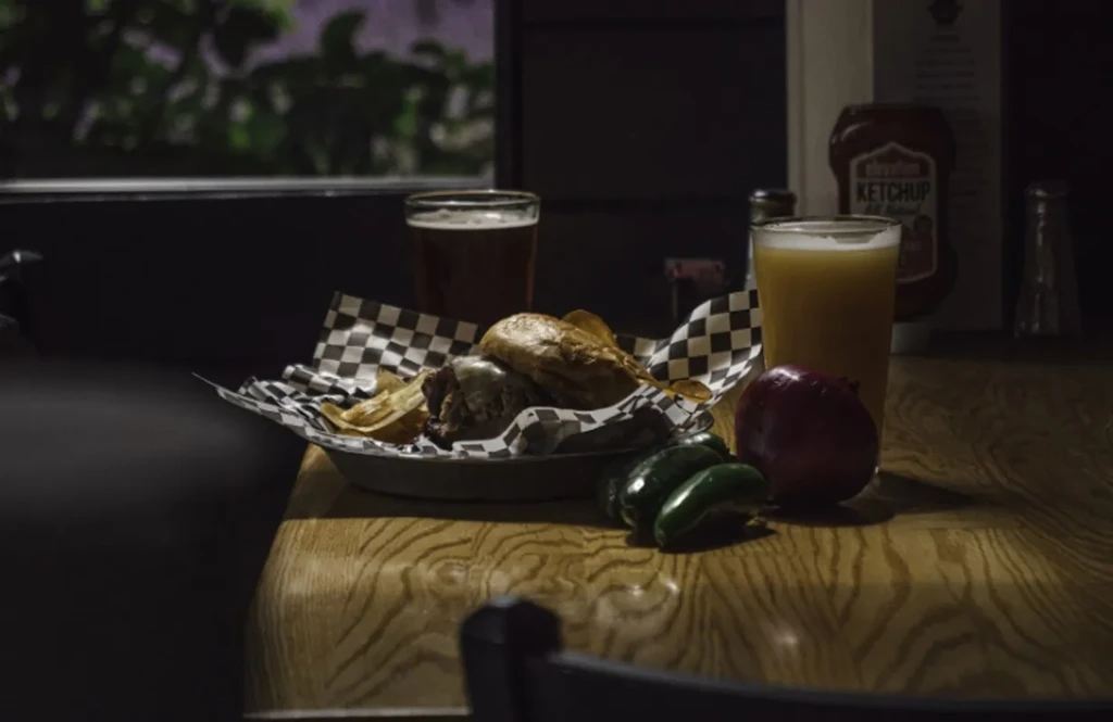 Dimly lit sandwich, chips, beers, onion, and jalapeños on a wooden table.