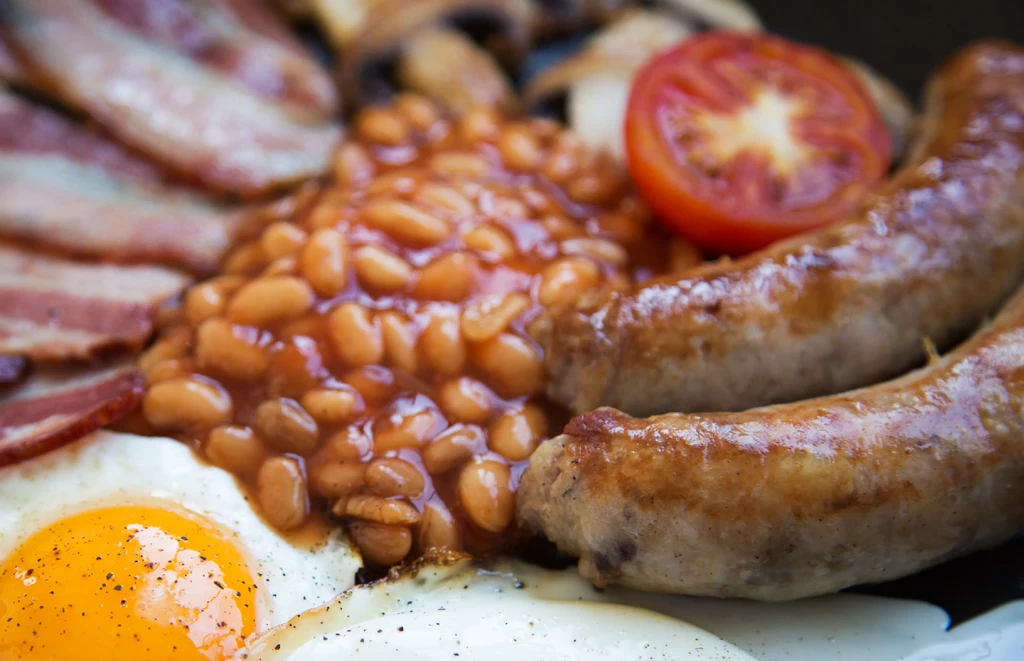 A plated full English breakfast featuring egg, beans, sausages, bacon, and mushrooms.