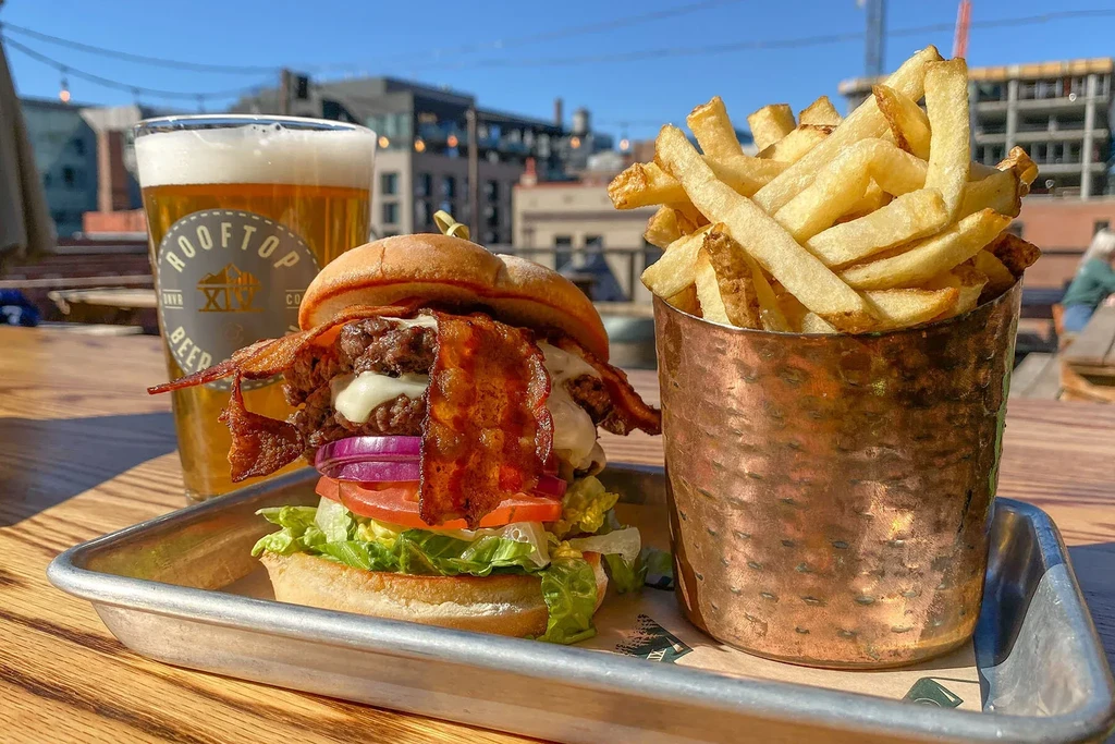 Close-up: gourmet burger, fries, and "ROOFTOP BEER CO" beer outdoors.