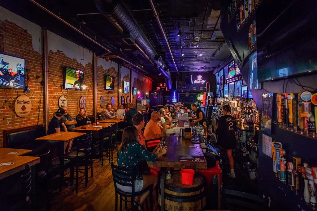 Bustling industrial bar interior with patrons, bartenders, TVs, and neon lights.