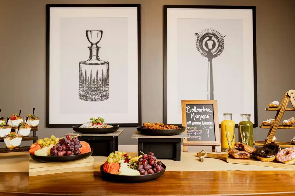 Warm-toned buffet spread with pastries, fruit, juices, and mimosas sign.