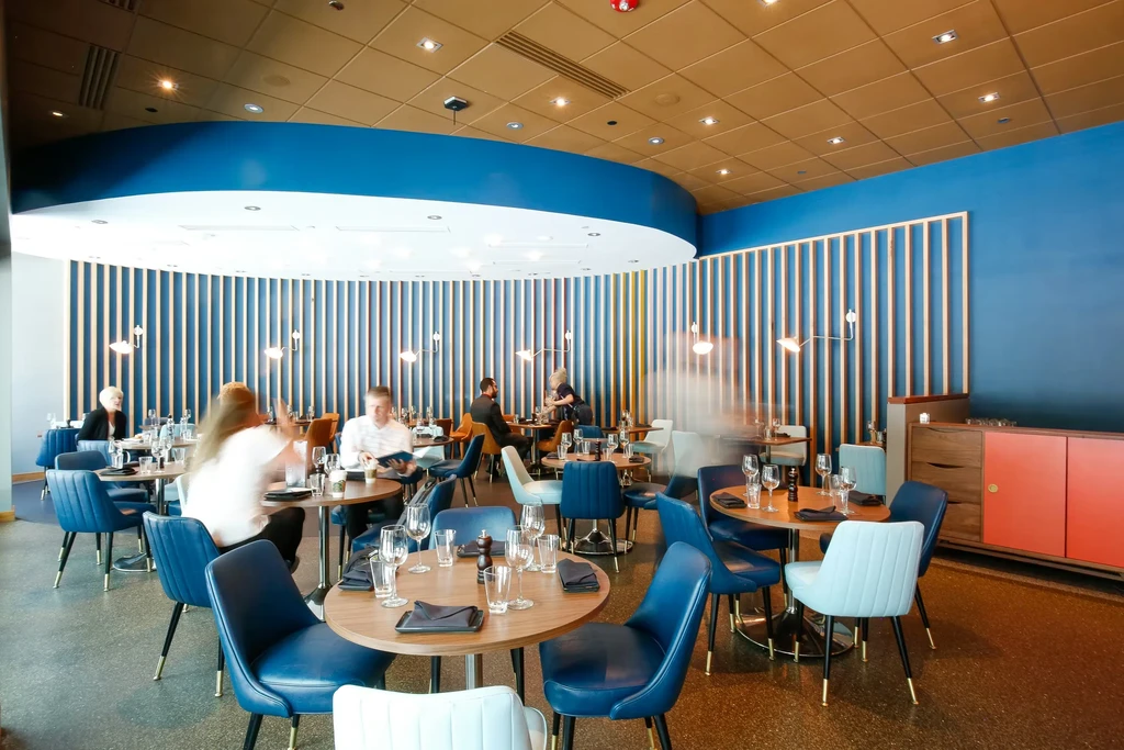 Modern restaurant interior with blue walls, wood ceiling, set tables, and blurred patrons.