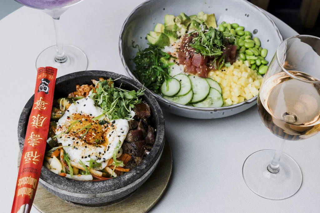 Overhead shot of bibimbap and poke bowls with a cocktail and rosé wine.