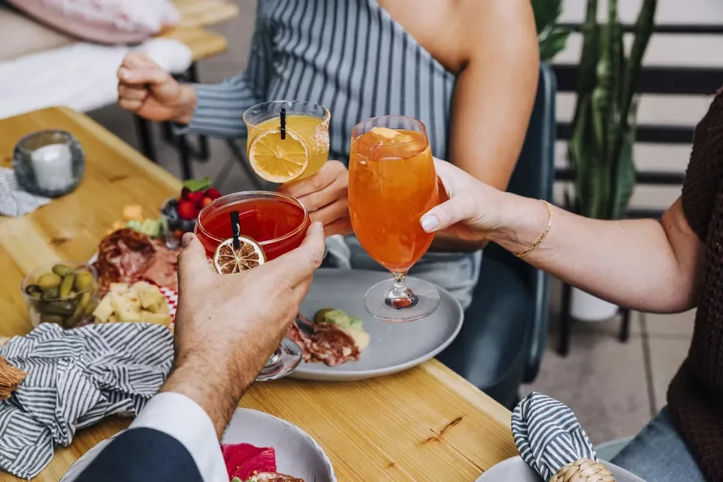 Hands raise three colorful cocktails above charcuterie and appetizers.