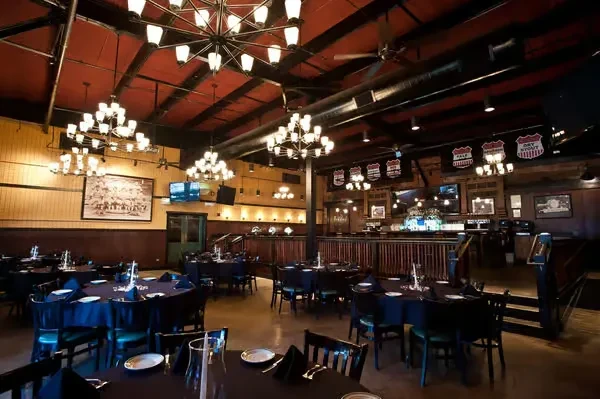 Empty, spacious restaurant with dark tables, exposed ducts, and blue-lit bar.