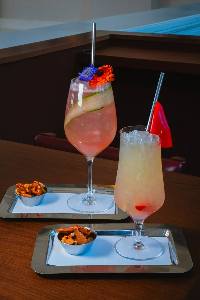 Two colorful, elaborately garnished cocktails with snacks on silver trays.