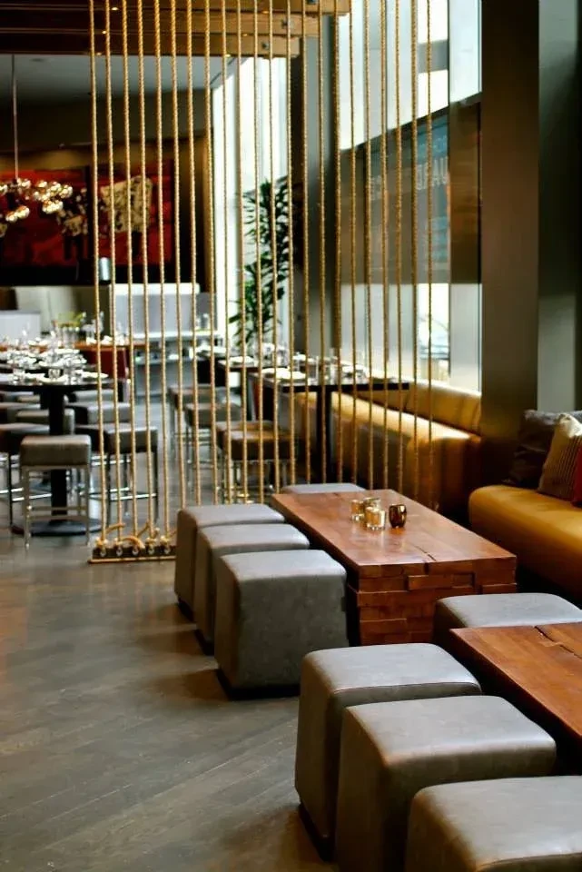 Modern restaurant lounge with golden rope divider, grey ottomans, and dining area.