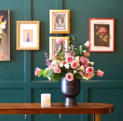 Large pink floral arrangement, candle, framed art, dark green wall.