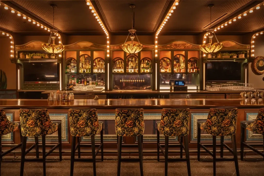 Warmly lit bar with wooden counter, floral stools, and illuminated bottles.