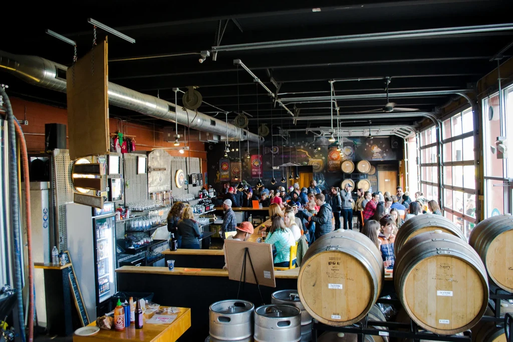 Bustling industrial brewery interior with barrels and many patrons.