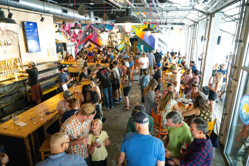 Busy modern brewery/bar with patrons, colorful murals, and open doors.