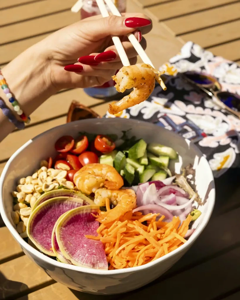Hand with chopsticks picks shrimp from colorful outdoor poke bowl.