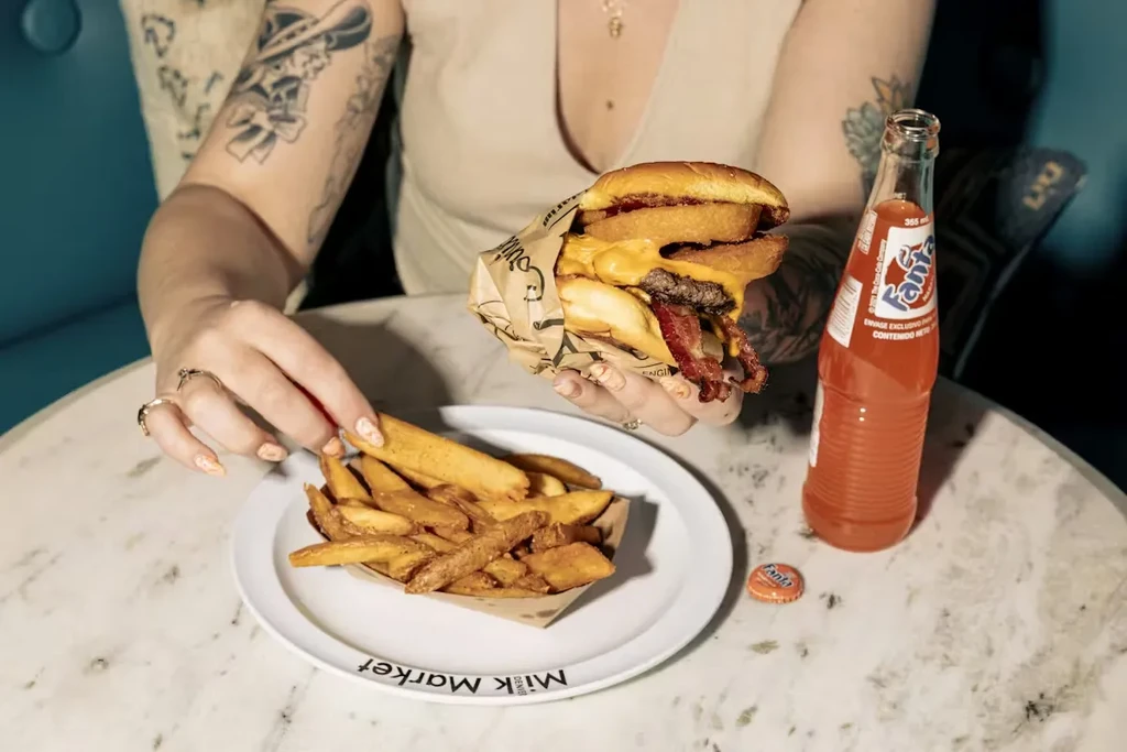 Tattooed hands hold a large burger, fries, and orange Fanta.