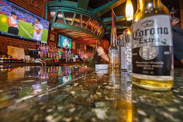 Reflective bar counter with Corona, liquor bottles, and TVs showing sports.