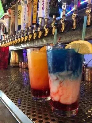 Two vibrant layered cocktails on a bar counter with beer taps.